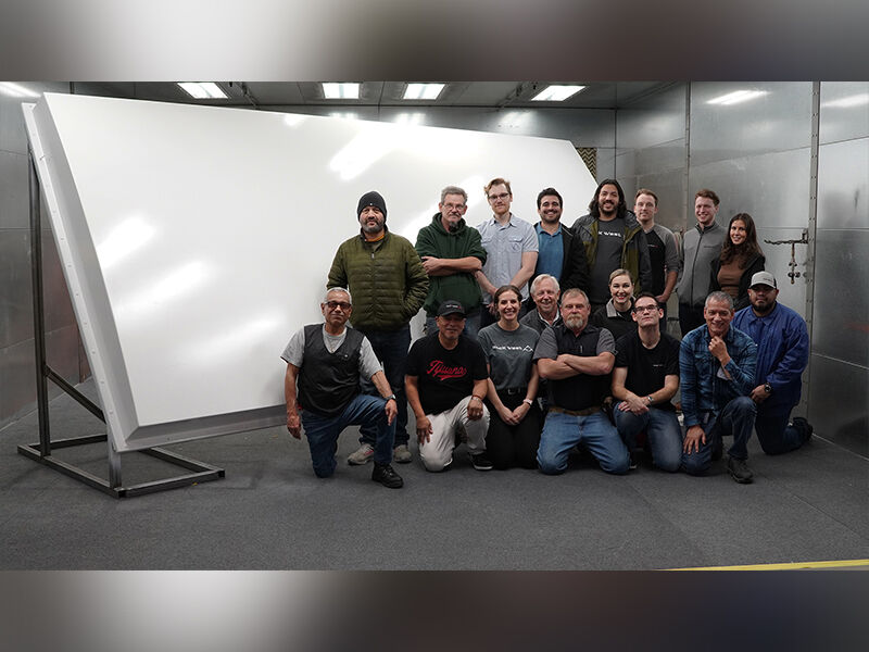 Image shows the radome tilted on a metallic stand in a large paint booth. The multiband large ground-based radome is painted white and in the center of the booth at an angle. The team who worked on the program is gathered in front and to the left side of the radome. 